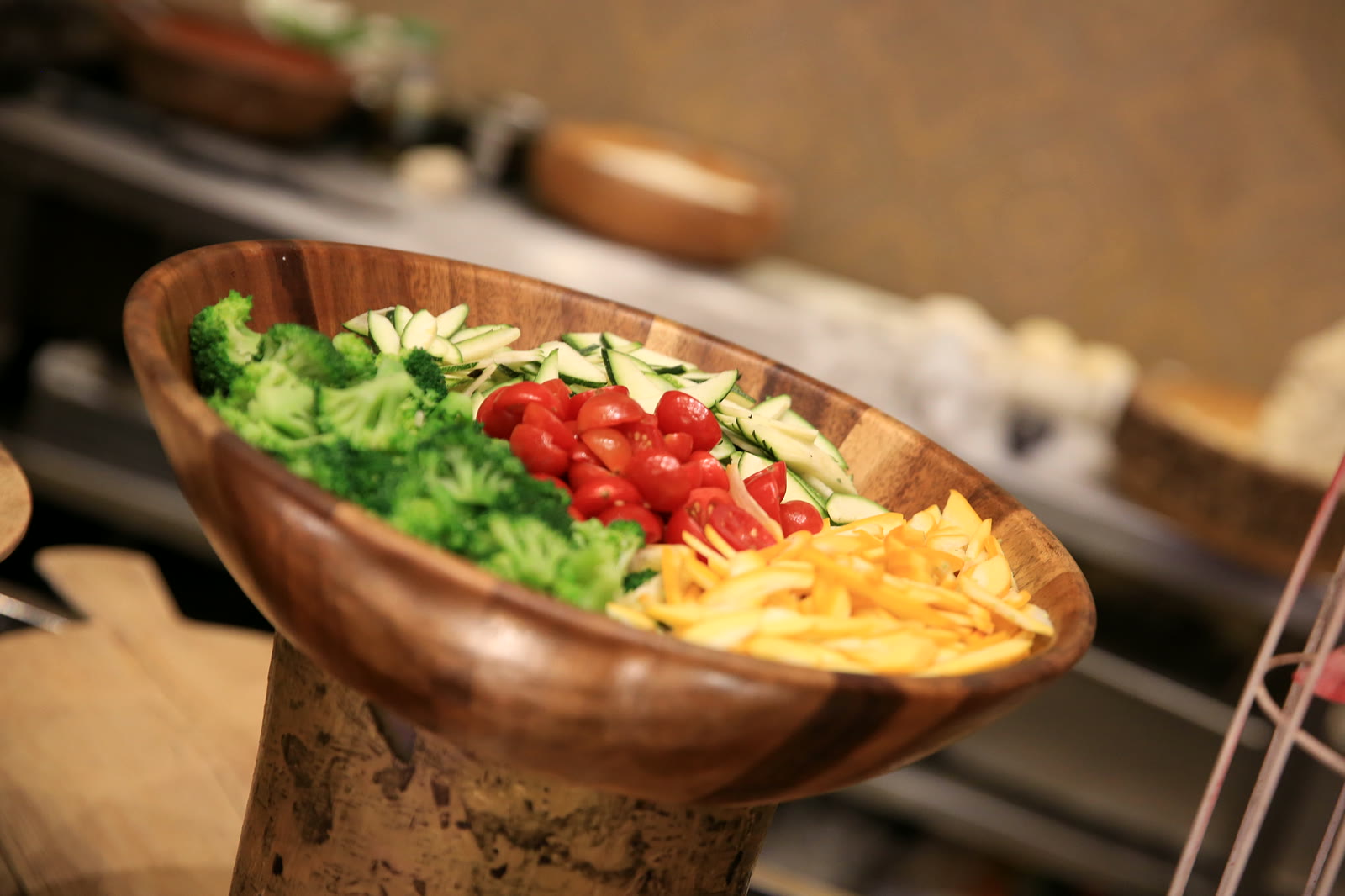 A wooden bowl of broccoli, cucumber, cherry tomatoes and carrot on a carved stand